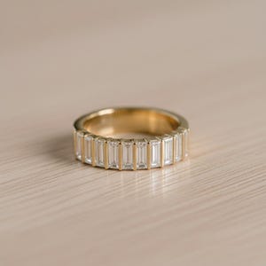 May include: A gold band ring featuring a series of rectangular, clear gemstones set around the circumference. The ring is set on a light-colored wooden surface, with the ring's gold band reflecting the light.
