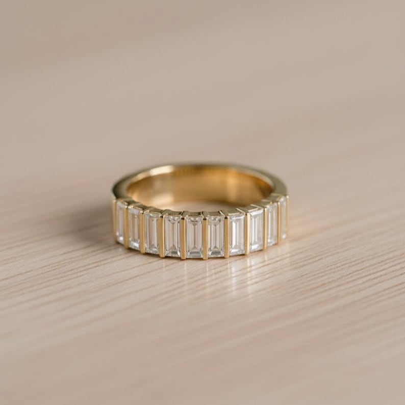 May include: A gold band ring featuring a series of rectangular, clear gemstones set around the circumference. The ring is set on a light-colored wooden surface, with the ring's gold band reflecting the light.