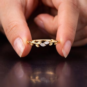 May include: A gold ring featuring a band designed with small metal leaves and two marquise-cut clear stones set in the center. The band has a polished finish and the leaf motifs extend along the sides of the ring, framing the two central stones.