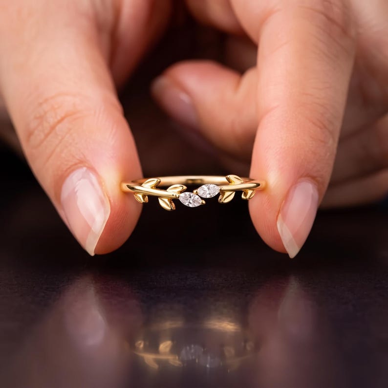 May include: A gold ring featuring a band designed with small metal leaves and two marquise-cut clear stones set in the center. The band has a polished finish and the leaf motifs extend along the sides of the ring, framing the two central stones.