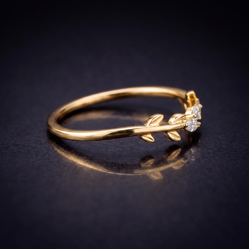 May include: A gold ring featuring a delicate band shaped like a vine with small leaves. The ring is accented with two small, clear, round-cut stones set within the leaf-like design, creating a nature-inspired look with a polished metallic finish.
