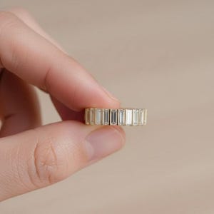 May include: A gold band ring with rectangular, clear gemstones set around the entire band. The ring is held between two fingers against a neutral background. The gemstones are evenly spaced and reflect light.