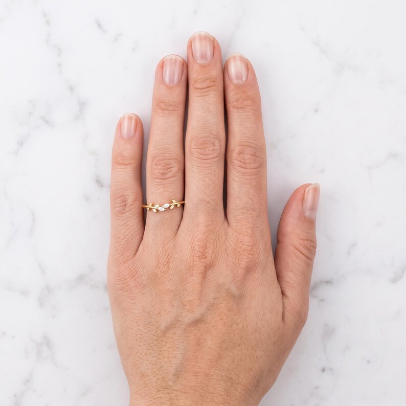 May include: A gold ring featuring a delicate branch design with small, marquise-cut white stones arranged like leaves along the band. The ring is worn on the ring finger, showcasing a slender, polished gold metal finish and a refined, nature-inspired aesthetic.