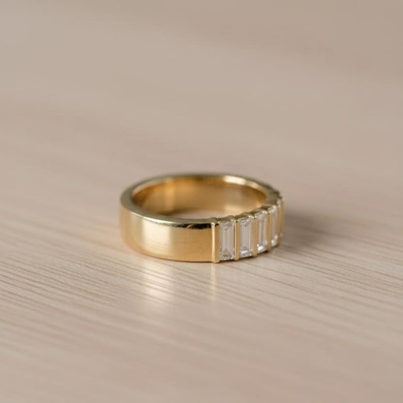 May include: A gold band ring with a row of rectangular, clear gemstones. The ring has a thick band and a polished finish. The gemstones are set in a line on the top of the ring.