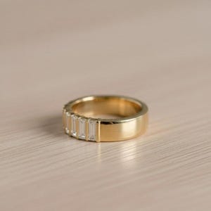 May include: A gold band ring with a row of rectangular, clear gemstones. The ring has a polished, smooth finish and is set against a light-colored wooden surface. The ring's design is simple and elegant.
