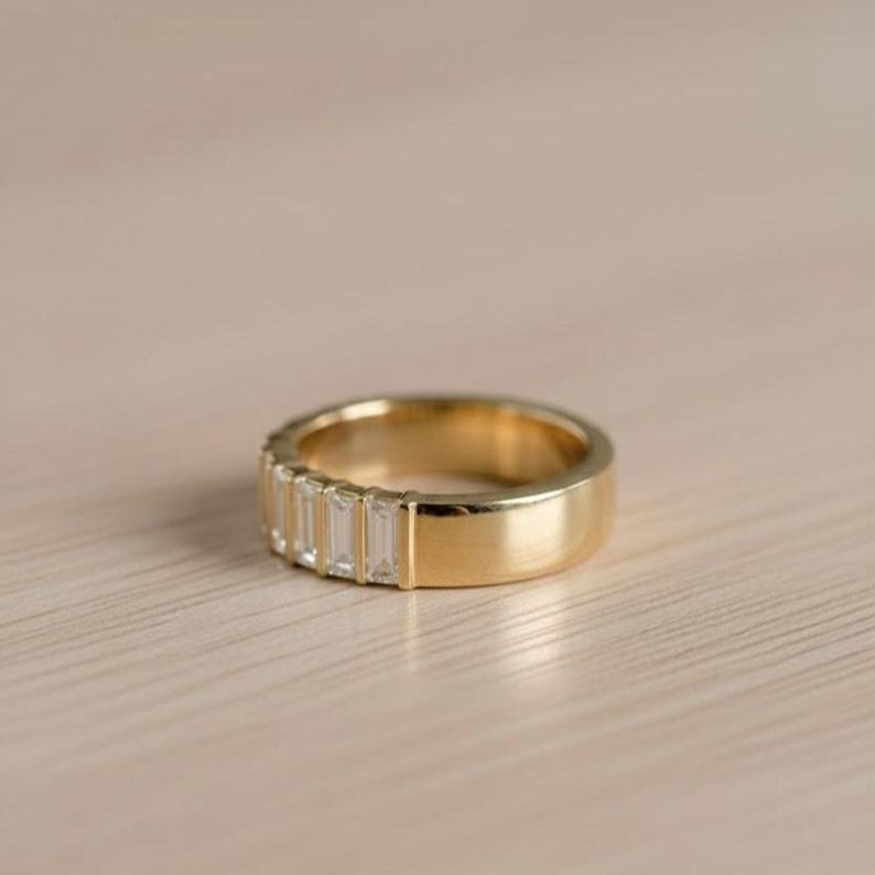 May include: A gold band ring with a row of rectangular, clear gemstones. The ring has a polished, smooth finish and is set against a light-colored wooden surface. The ring's design is simple and elegant.