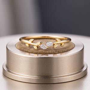 May include: A gold ring featuring a band shaped like a delicate vine with leaves. The center of the ring is open and accented with two small, marquise-cut clear stones positioned to resemble buds at the ends of the vine-like metal branches.