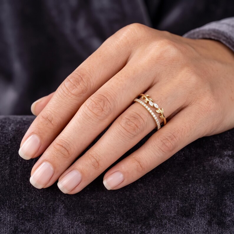 May include: A gold ring featuring two bands joined together. One band is a thin, plain gold wire adorned with small, marquise-cut clear stones arranged in a leaf-like pattern. The second band is a simple, slender gold wire set with tiny clear stones.