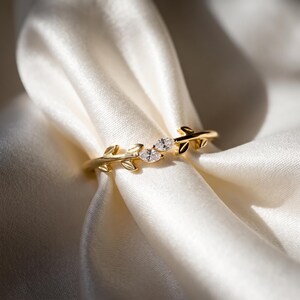 May include: A gold ring featuring a band designed with metallic leaf motifs and two marquise-cut clear stones set in the center. The band has a polished finish and the leaf shapes extend along the sides of the stones, creating a nature-inspired look.