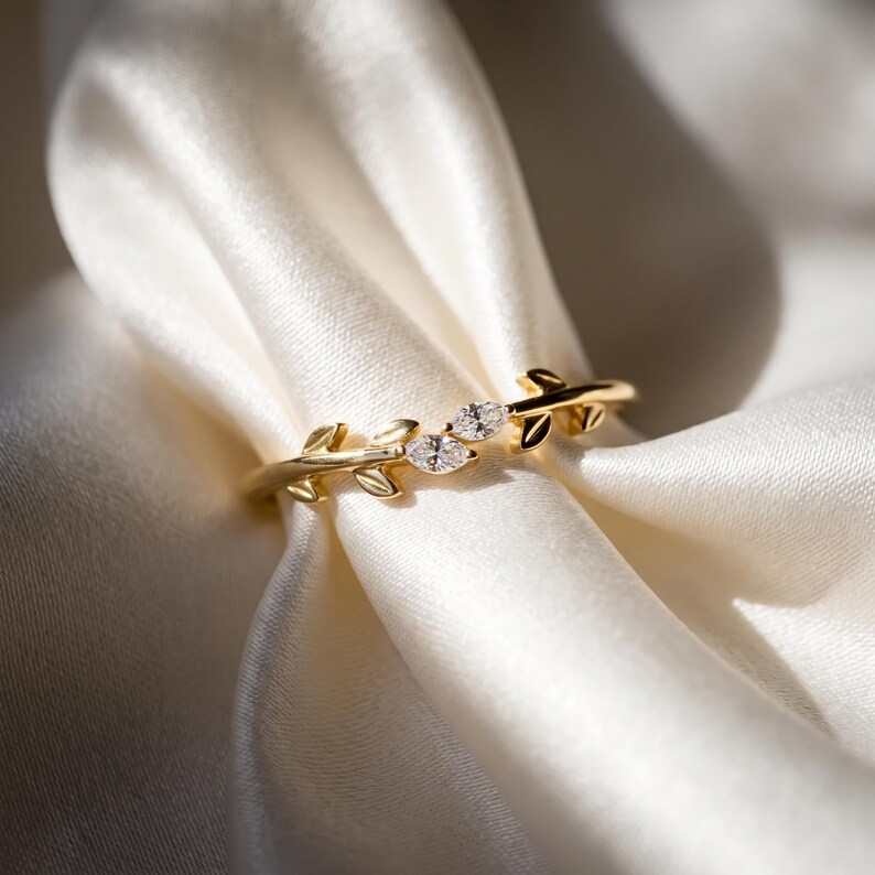 May include: A gold ring featuring a band designed with metallic leaf motifs and two marquise-cut clear stones set in the center. The band has a polished finish and the leaf shapes extend along the sides of the stones, creating a nature-inspired look.