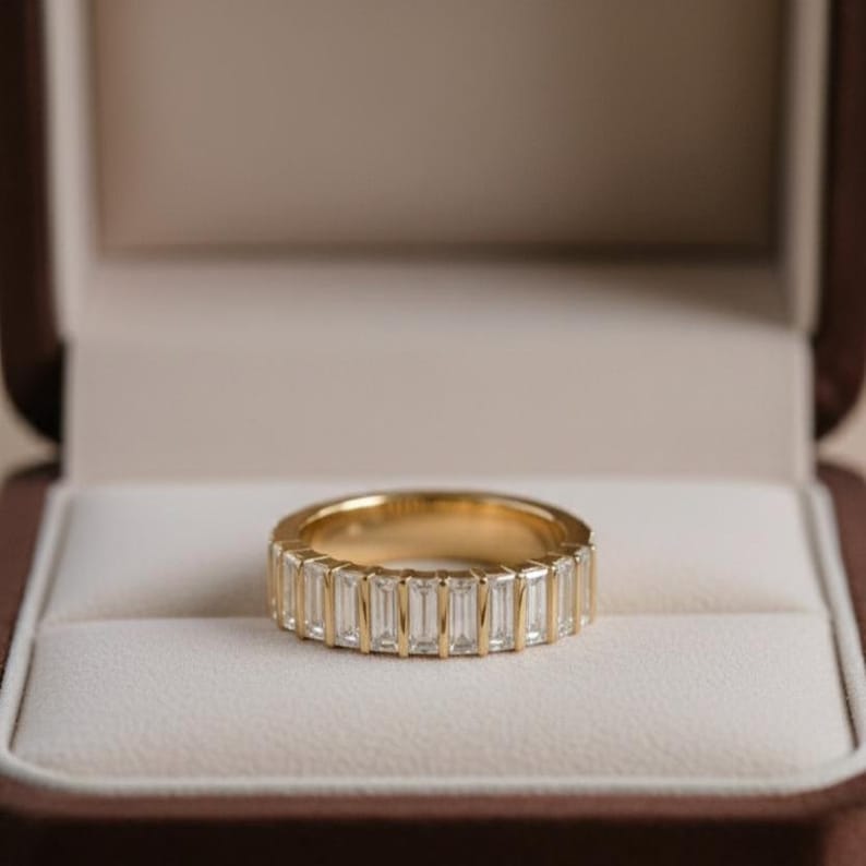 May include: A gold band ring with a row of rectangular, clear gemstones. The ring is set in a gold band and is displayed inside a brown jewelry box with a white interior.