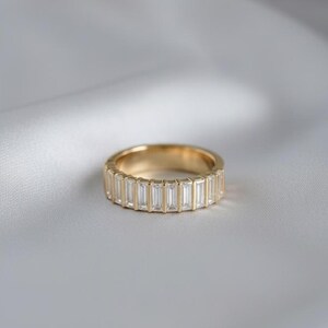 May include: A gold band ring with a continuous row of rectangular, clear gemstones. The ring is set against a soft, white background, highlighting the ring's polished gold and the sparkle of the stones.