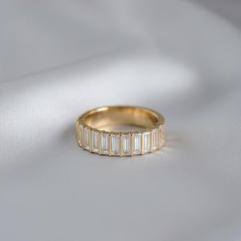 May include: A gold band ring with a continuous row of rectangular, clear gemstones. The ring is set against a soft, white background, highlighting the ring's polished gold and the sparkle of the stones.