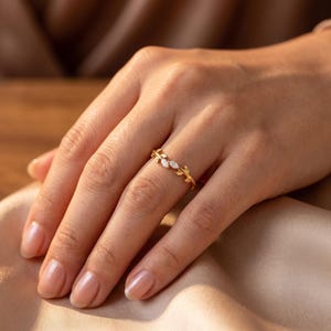 May include: A gold ring featuring a delicate vine-like band with small leaves and two marquise-cut clear stones set in the center. The metal has a polished finish, and the design wraps around the finger with a slender, organic aesthetic.