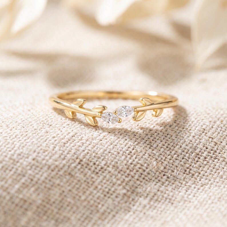 May include: A gold ring featuring a delicate branch design with small leaves and two marquise-cut clear stones set in the center. The band is thin and polished, showcasing a nature-inspired aesthetic with a bright, reflective metallic finish.