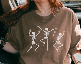 May include: A brown t-shirt featuring a white graphic of three dancing skeletons. The skeletons are in various poses, with arms and legs outstretched. The t-shirt is a solid brown color.