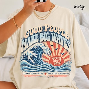 Può includere: Una t-shirt color avorio con una grafica che riporta il testo "GOOD PEOPLE MAKE BIG WAVES" in un carattere retrò. Il design include una grande onda blu, un sole rosso, tre uccelli in volo e il testo "VOTE NOV 3 2026" e "CLEANER GOVERNMENT BRIGHTER TOMORROWS".