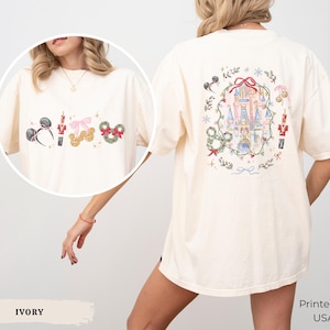 May include: An ivory t-shirt featuring a back graphic of a castle surrounded by a holiday wreath, nutcrackers, and mouse ears. The front chest area displays a smaller print of mouse ears, a nutcracker, and a bow. The shirt is printed in the USA.
