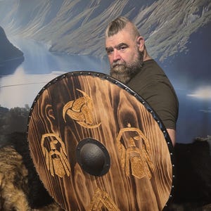 May include: A round wooden shield with a dark brown finish and intricate gold-colored carvings of Viking symbols. The shield has a black metal boss in the center and a black metal rim. The shield is held by a person wearing a green shirt.