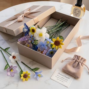 May include: A beige gift box with a bouquet of colorful glass flowers, including sunflowers, daffodils, and other blooms. The box is tied with a satin ribbon. A small velvet pouch and a card with text are also present.