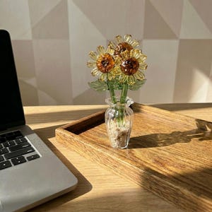 May include: A glass vase holding three glass sunflowers with yellow petals and brown centers. The vase sits on a wooden tray, next to a laptop on a wooden desk. The sunflowers have green leaves.