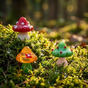 May include: Three small glass mushroom figurines nestled in green moss. One mushroom has a red cap with white spots, one is orange with a red flower detail, and the third is green with small red dots. Each piece features a smooth, glossy finish.