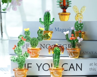 May include: A collection of glass cactus figurines in various shapes and colors, each potted in a small amber-colored container. Some cacti have colorful flower accents. The figurines are arranged on top of books with the text "IF YOU CAN".
