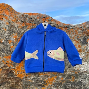 May include: A blue hooded fleece jacket with a zipper closure and two fish appliques on the front. The fish are made of fabric scraps in shades of green, brown, and pink.