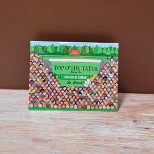 May include: A colorful illustration of a food product box. The box features the text "TOP O' THE TATER" and "Onion & Chive" with the phrase "So Good!" The artwork depicts a crowd of people around a food container.