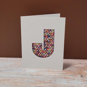 May include: A folded white greeting card featuring a large, colorful letter "J". The letter is filled with a pattern of small, stylized faces in various colors. The card is set against a brown background and a wooden surface.