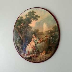 May include: An oval wall plaque featuring a pastoral scene with two figures in period clothing, one playing a lute. The artwork includes a fountain, lush trees, a small dog, and a basket of flowers, all enclosed within a dark red frame.
