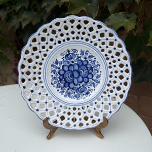May include: A round ceramic plate featuring a white center with a blue floral pattern and a scalloped, lattice-style openwork border decorated with small blue dots. The plate has a wavy edge and is displayed on a small, brown stand.