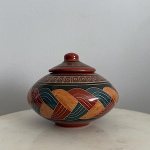 May include: A round, lidded ceramic vessel featuring a wide, squat body and a small knobbed lid. The surface is decorated with a repeating braided pattern in shades of terracotta, teal, and tan, accented by thin white lines and a geometric border.