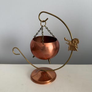 May include: A copper-colored metal cauldron with three small feet, suspended by three thin chains from a curved brass-toned stand. The stand features a decorative metal emblem of a spinning wheel attached to the side, resting on a circular base.