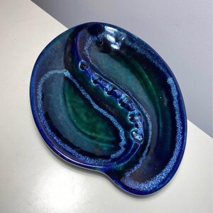 May include: An oval ceramic dish featuring a deep blue and emerald green glazed finish. The surface has a central raised ridge with indentations, creating a divided basin effect with a glossy, mottled texture throughout the piece.