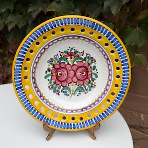 May include: A round ceramic plate featuring a white center with a hand-painted floral motif in shades of red, green, and purple. The wide yellow rim is decorated with a blue striped border and a series of evenly spaced circular cutouts.