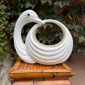 May include: A white ceramic swan-shaped planter featuring a ribbed, textured surface and a single dark eye. The swan's neck curves gracefully to form a circular opening, creating a hollow, rounded body designed to hold small items or plants.