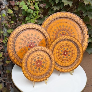 May include: A set of three round wooden decorative plates featuring a warm yellow finish and intricate hand-painted floral patterns in red, green, and brown. Each plate displays a central flower motif surrounded by concentric circular designs.
