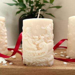 May include: Ivory-colored pillar candle with a detailed relief carving of Santa Claus in a sleigh pulled by reindeer. A red ribbon and white wax beads are on the wooden surface. The candle is unscented and has a central wick.