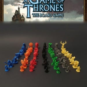 以下が含まれることがあります： 「A Game of Thrones」ボードゲーム、第2版。さまざまな色のゲームピースがあります。ゲームピースは青、オレンジ、赤、黒、緑、グレー、黄色です。ゲームのタイトルが画像の上部に表示されています。