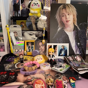 May include: A collection of BTS merchandise, including keychains, plush toys, bracelets, and photo cards. The items feature images of band members and the group's logo. The color palette includes yellow, pink, purple, and black.