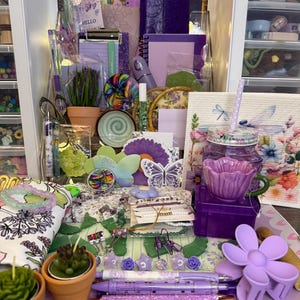 Puede incluir: Una colección de artículos de papelería, accesorios y objetos decorativos de color púrpura y lavanda. Incluye bolígrafos, clips, pinzas para el pelo con forma de flor, pulseras y pequeñas plantas en macetas. También se ve una variedad de cuadernos, diarios y papel decorativo.