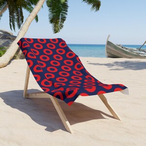 May include: A wooden beach chair with a navy blue and red patterned towel draped over it. The towel features a repeating design of red ovals on a navy blue background. The chair is on a sandy beach with a boat in the background.