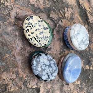May include: Four polished gemstone stones in oval shapes. One stone is cream-colored with black spots, another is white with gray veining, a third is black with white snowflake patterns, and the last is blue.