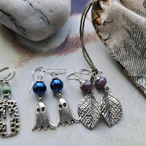 May include: Three pairs of earrings with different designs. One pair has green beads and cactus-shaped charms. Another features blue beads and ghost charms. The third pair has purple beads and leaf charms. A small fabric pouch is also present.