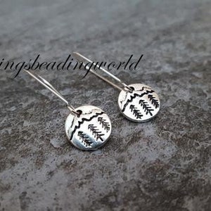 May include: Silver earrings with a small, round charm featuring a stylized mountain scene with trees. The earrings are on a gray textured surface.