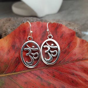 May include: Silver earrings with a circular design featuring the Om symbol. The earrings are hanging from a red leaf.