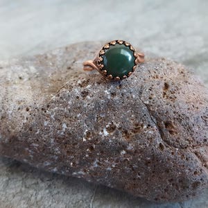 May include: A copper ring with a green gemstone set in a decorative bezel. The ring is resting on a rough, brown rock.
