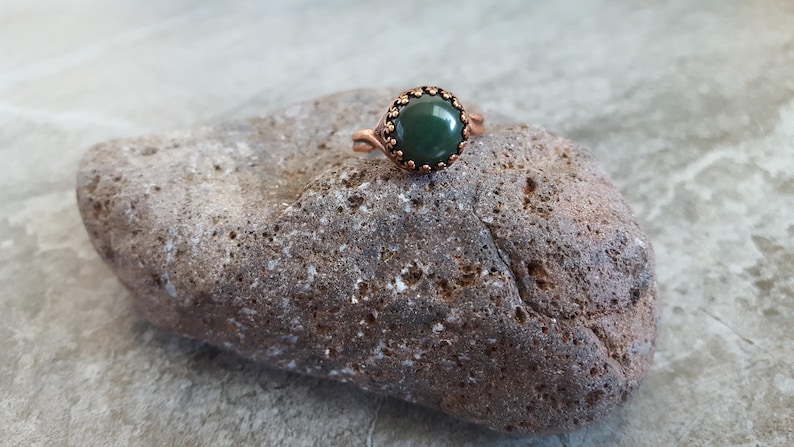 May include: A copper ring with a green gemstone set in a decorative bezel. The ring is resting on a rough, brown rock.