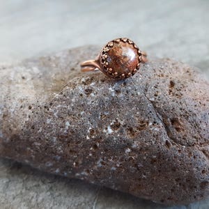 May include: A copper ring with a brown and orange stone set in a copper bezel. The ring is sitting on a rough, gray rock.